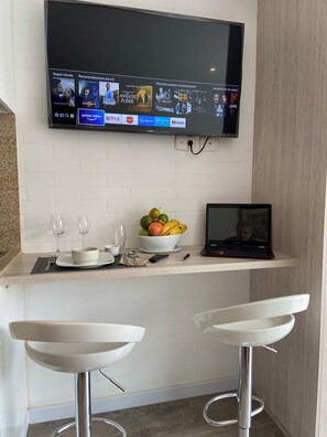 Une cuisine moderne avec un téléviseur à écran plat fixé au mur, qui affiche l’interface d’un service de diffusion en continu. On y trouve deux tabourets de bar blancs et un ordinateur portable sur un comptoir en bois.