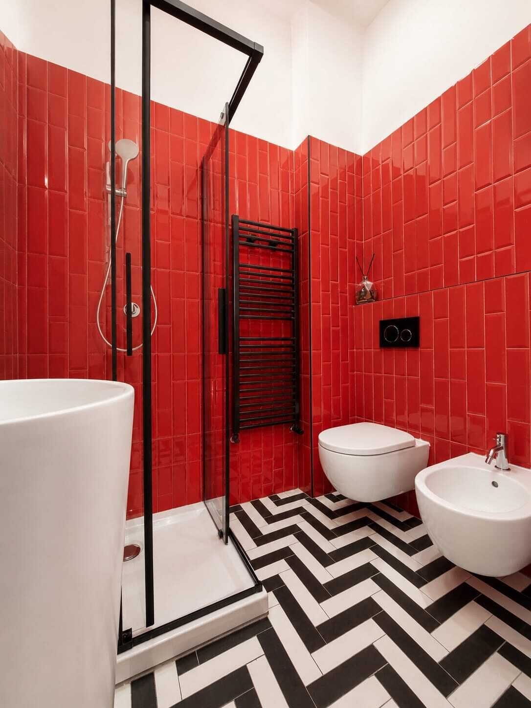 Un baño con paredes de azulejos rojos, suelo a cuadros blancos y negros tipo chevron, cabina de ducha de vidrio, inodoro blanco y lavamanos blanco con bidé.
