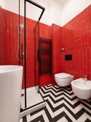 A bathroom with red tiled walls, a black and white chevron floor pattern, a glass shower enclosure, a white toilet, and a white sink with a bidet. - La Presentosa Studio Apartment (Bologna)