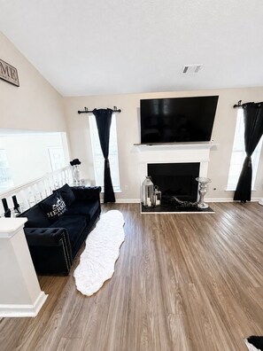 A living room with a black sofa, a white fireplace, and a flat-screen TV mounted above it. - Charming 3-bedroom house with AC in pleasant Riverdale (Riverdale)