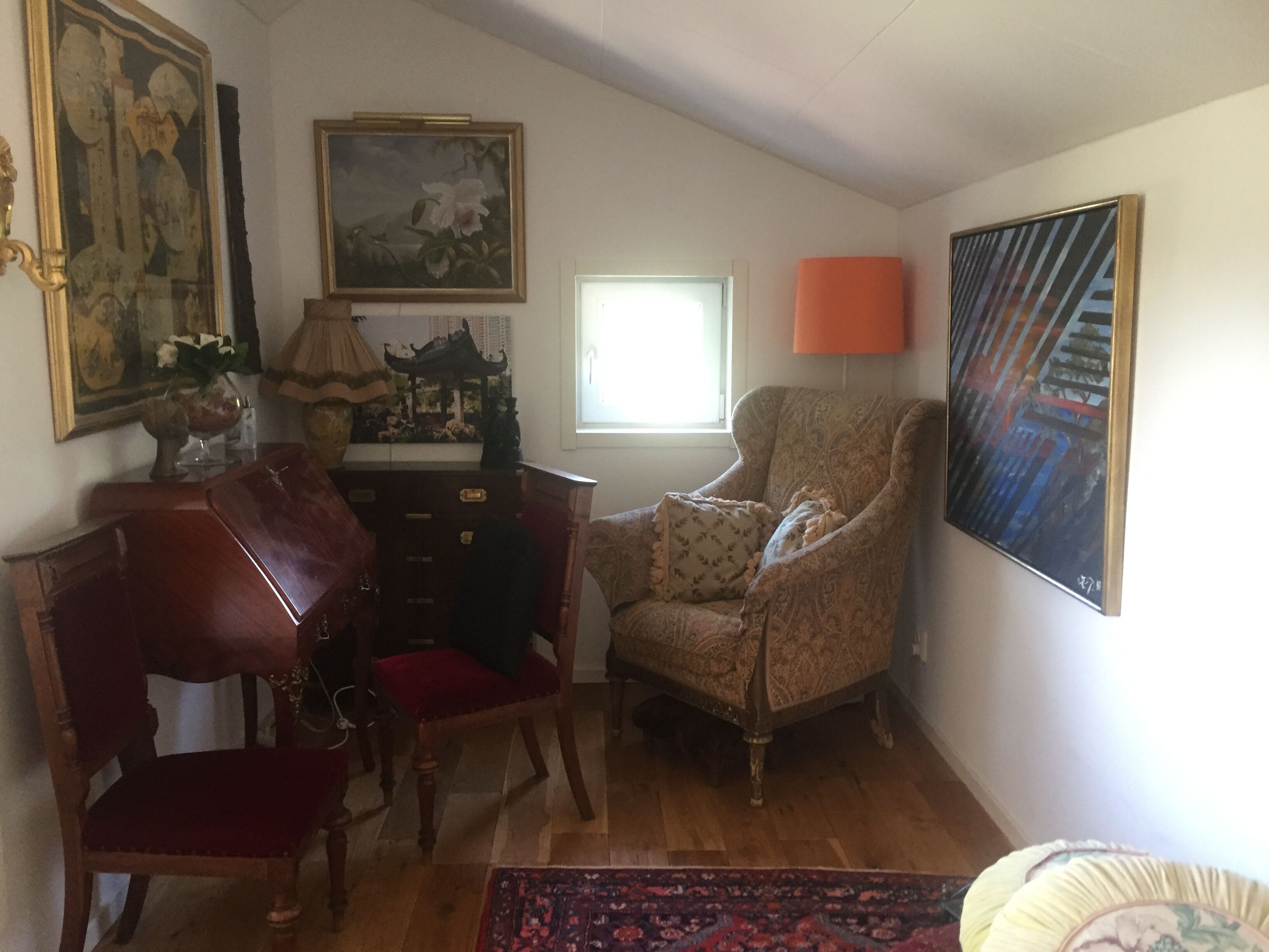 A room with a wooden chair, a desk, and a lamp. There is a window with a view of the outside.