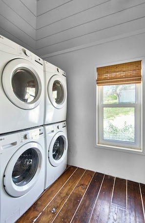 A laundry room with four stacked front-loading washing machines, a window with a view of greenery, and wooden flooring. - Bohemia Bunkhouse - Steps to Pink Sand, Heated Pool (Governor's Harbour)
