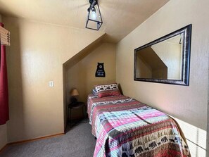 A bedroom with a sloped ceiling, a bed with a patterned bedspread, a nightstand, a wall clock, and a mirror. - Northern Escape | Sauna, Dock & Lake Access (Butternut)