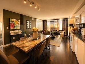 A modern kitchen with a dining area, a living room with a sofa and a coffee table, and a large window with curtains.