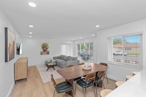 Una sala moderna con un sofá gris, una mesa de comedor con sillas, un aparador de madera y un ventanal con vista a la calle.
