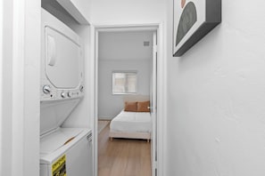 Un pasillo estrecho que conduce a un dormitorio con una cama, una ventana con persianas y un cuadro colgado en la pared.