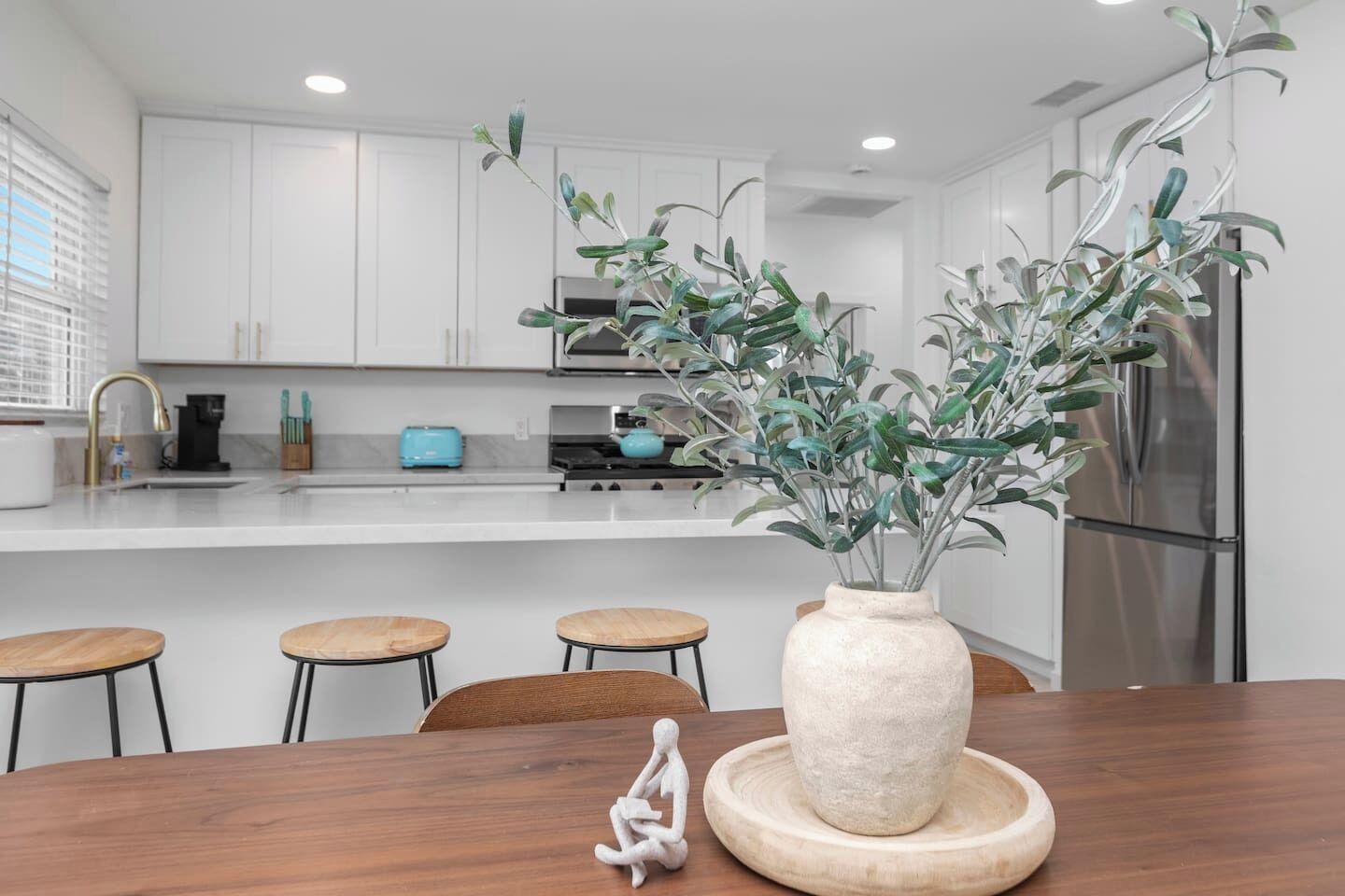 A modern kitchen with white cabinets, a stainless steel refrigerator, a blue toaster, and a vase with green plants on the island.