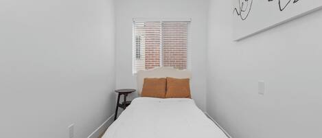 Un dormitorio con cama, una mesita, una ventana con persianas y una pared con un letrero.