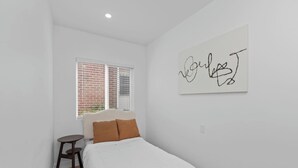 Une chambre à coucher avec un lit, une petite table, une fenêtre avec des stores et un mur orné d’une œuvre d’art abstraite.