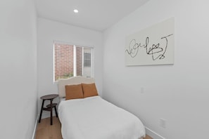 Un dormitorio con una cama, una mesita, un ventanal con persianas y una pared decorada con una obra de arte abstracta.