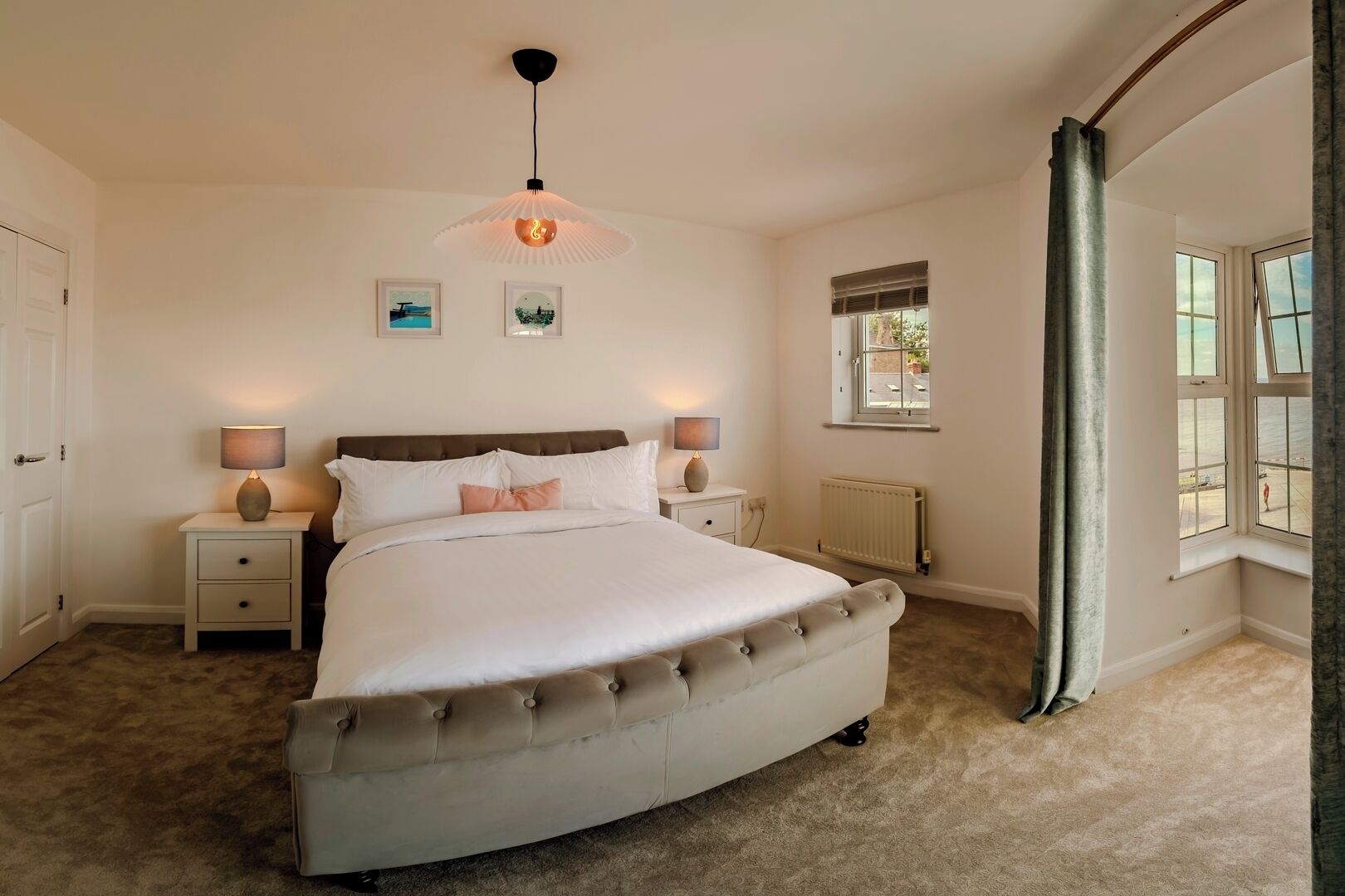 A bedroom with a large bed, two bedside tables with lamps, a radiator, and a window with a view.