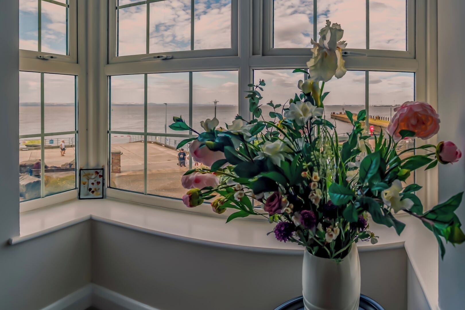 Een kamer met een groot raam dat uitkijkt op een strand, een vaas met bloemen en een lijst met een foto op de vensterbank.