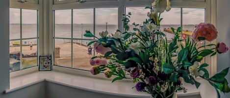 Una habitación con un ventanal grande con vistas a una playa, un florero con flores y un cuadro en el alféizar de la ventana.