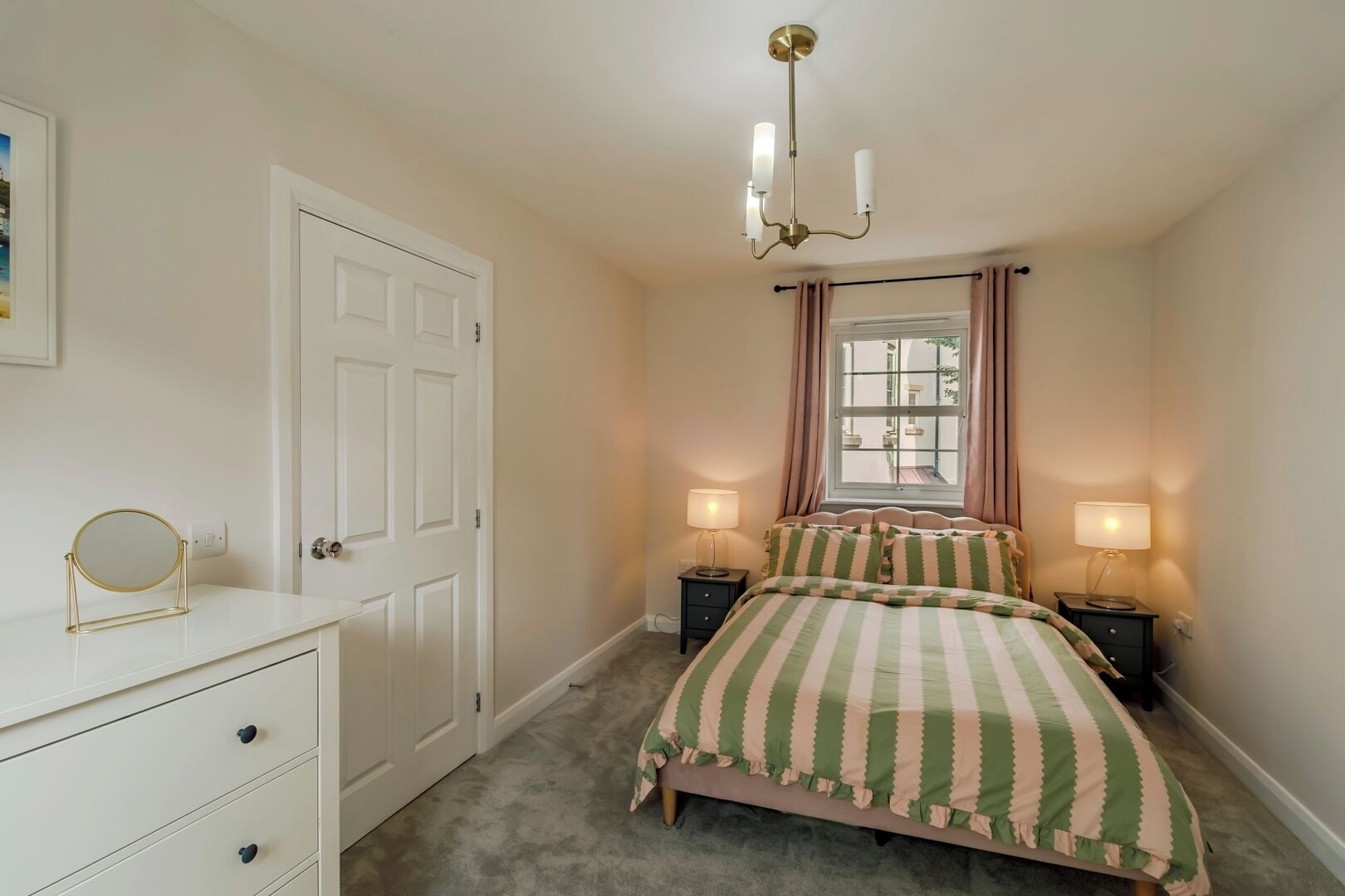 A bedroom with a bed, bedside tables, a dresser, a window with curtains, and a chandelier.