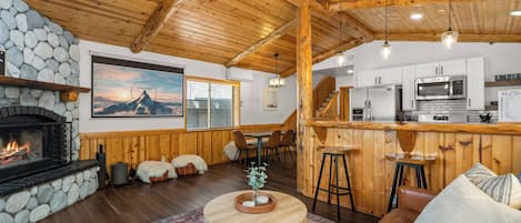 A cozy interior with a stone fireplace, wooden ceiling, a bar area, and a kitchen.