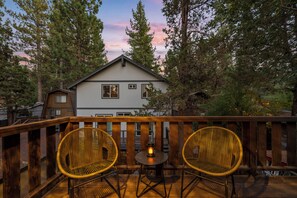 A wooden deck with two wicker chairs and a small table, overlooking a house surrounded by trees. - Cozy modern cabin in Lower Moonridge, 5 minutes from Big Bear Mountain! (Big Bear Lake)