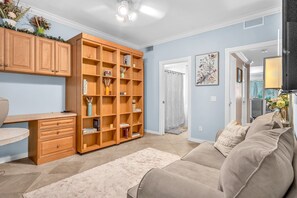 Ein Wohnzimmer mit einem beige gefärbten Sofa, Holzregalen, einem Schreibtisch und einem großen Fenster.