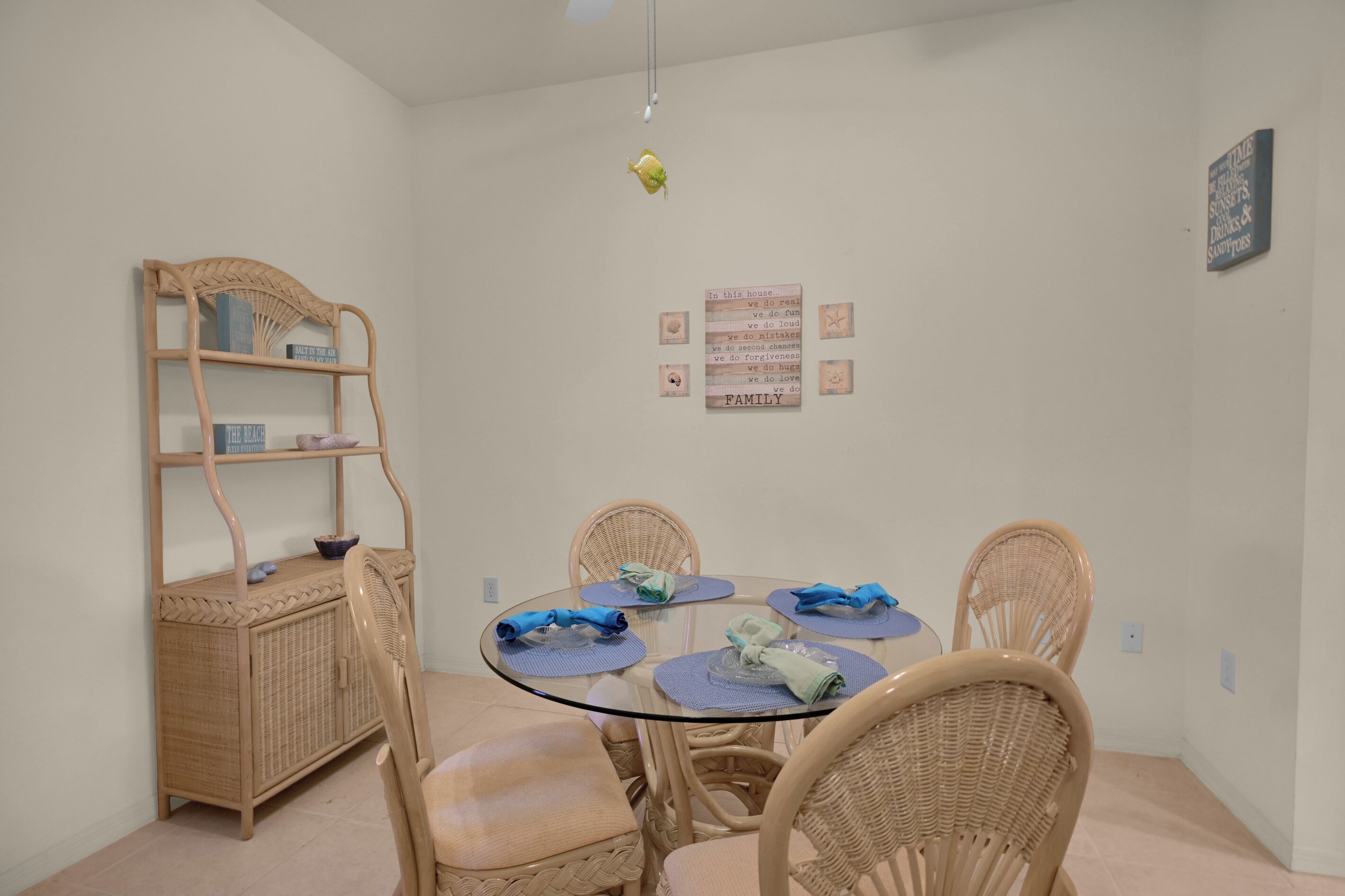 Uma área de refeições com uma mesa redonda de vidro, cadeiras de vime e um aparador de vime com livros.