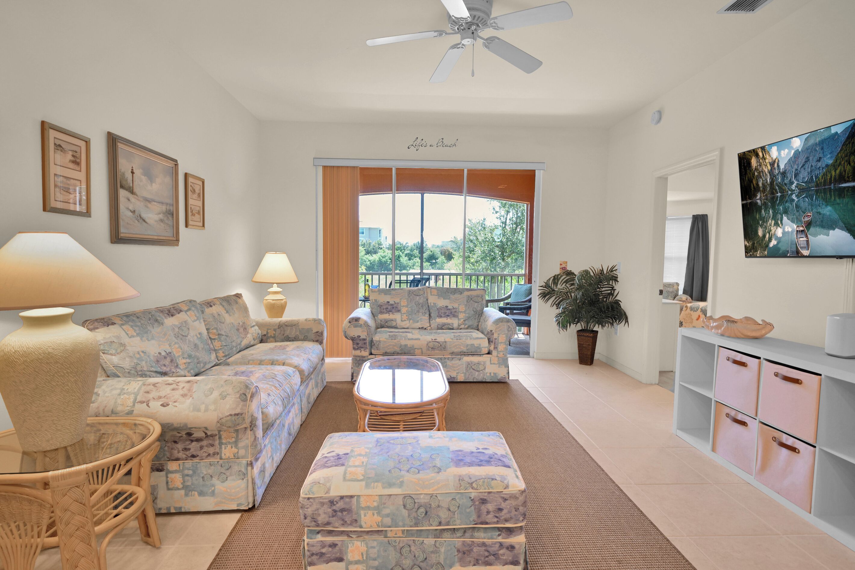 A living room with a sofa, armchairs, a coffee table, and a television. There is a balcony with a view of the outdoors.