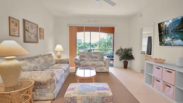 A living room with a sofa, armchairs, a coffee table, and a television. There is a balcony with a view of the outdoors.