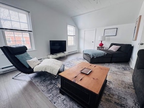 A modern living room with a black leather chair, a wooden coffee table, a grey sofa, and a television.