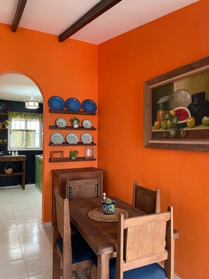A dining area with a wooden table and chairs, a wall with decorative plates, and a painting of fruits. - The Mexican Casita (Cancun)
