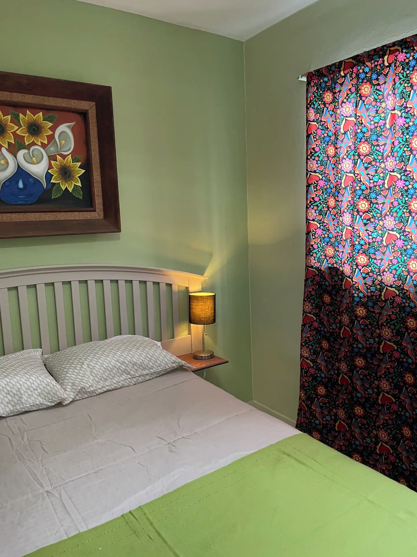 A bedroom with a bed, a nightstand with a lamp, a painting on the wall, and a patterned curtain.