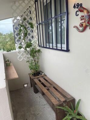 Een balkon met een houten bankje, een plant in een pot, een decoratieve metalen poort en een aangebracht huisnummer.