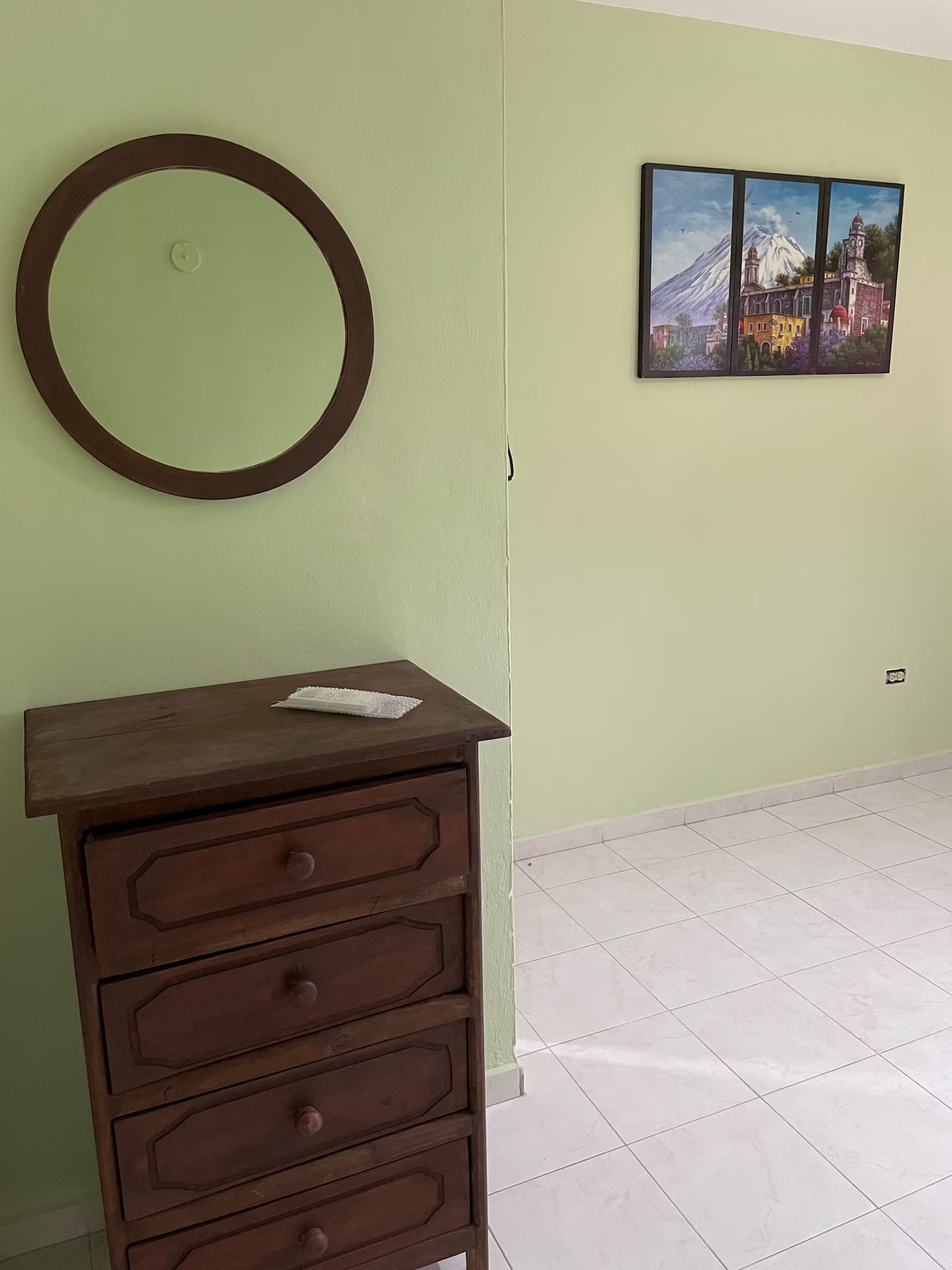 A room with a wooden dresser, a round mirror, and a wall-mounted painting.