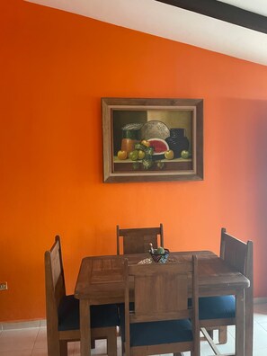 Een eethoek met een houten tafel en stoelen, een lijstwerk schilderij erboven en een oranje wand.