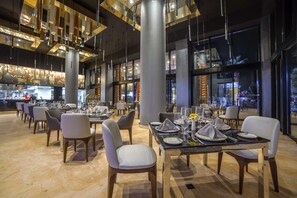 Un interior de restaurante moderno con techos altos, luces colgantes y una zona de bar al fondo.