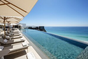 Zona junto a la piscina con sillones de descanso y sombrillas, con vistas al océano azul y despejado.