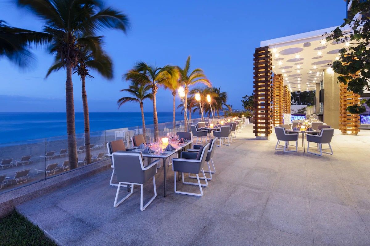 An outdoor dining area with tables and chairs, palm trees, and a view of the ocean.