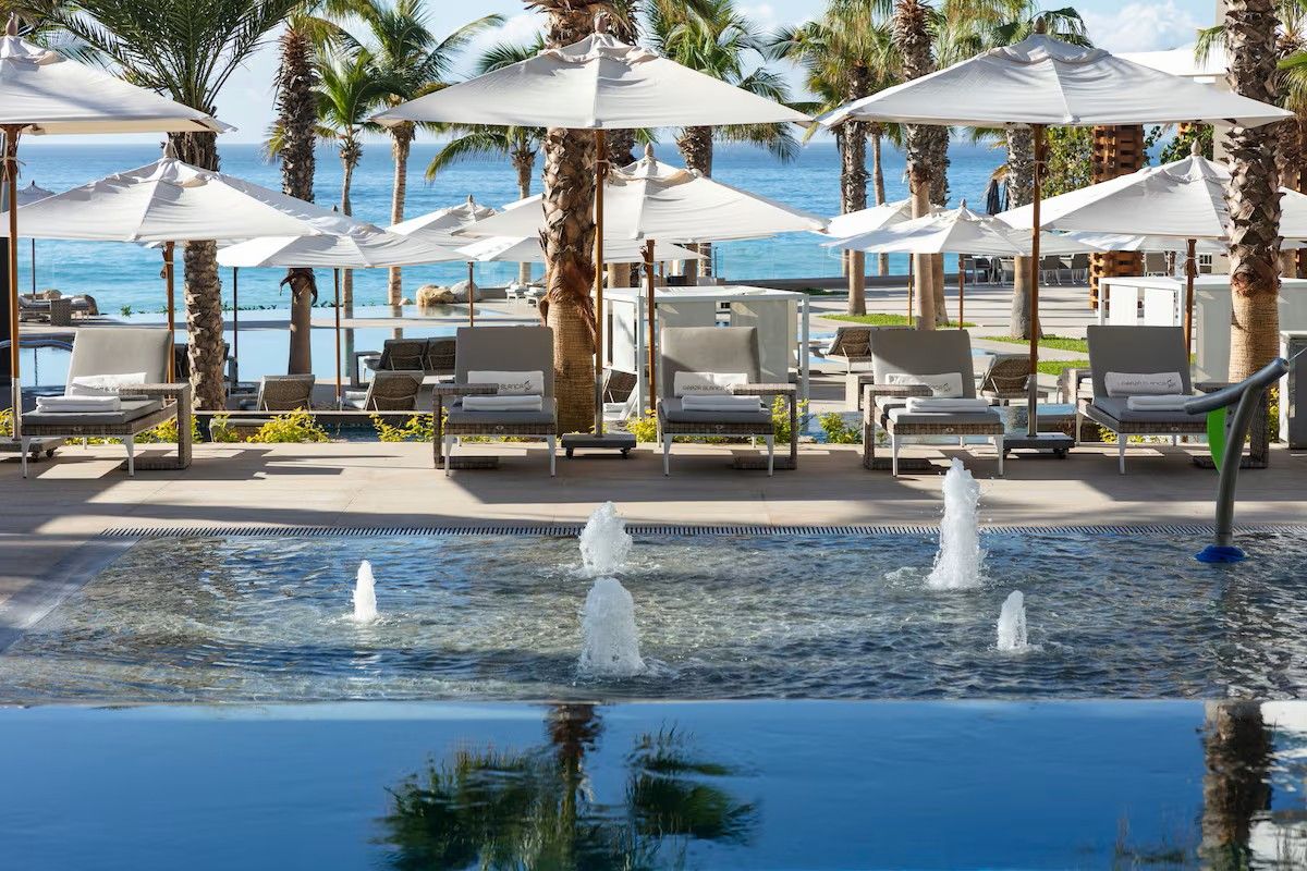 A pool area with lounge chairs and umbrellas, palm trees, and a view of the ocean.