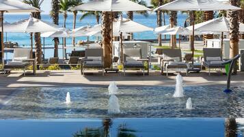A pool area with lounge chairs and umbrellas, palm trees, and a view of the ocean.