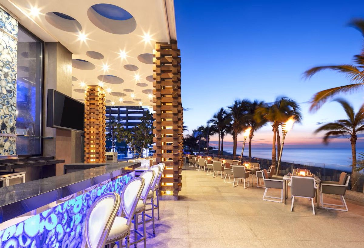 A bar area with a view of the ocean, outdoor seating, and a modern architectural design with circular light fixtures.