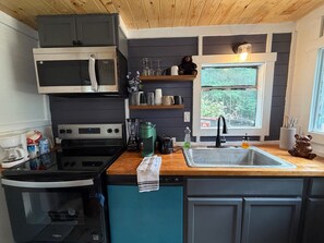 A compact kitchen with a microwave, stove, and sink. There are shelves above the counter holding various items.
