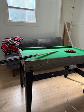 Een biljart met een knuppel en ballen, een bank met een gestreepte deken, en een raam met uitzicht.