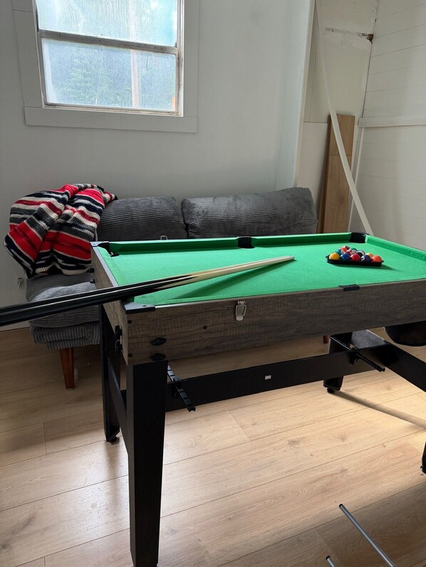 A pool table with a cue stick and balls, a sofa with a striped blanket, and a window with a view.