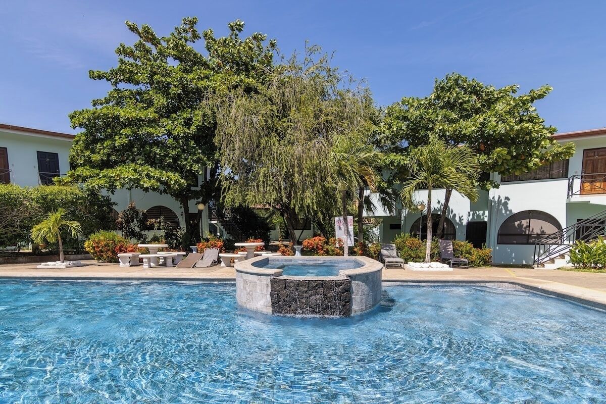 A swimming pool with a central water feature, surrounded by lush greenery and a building with arched windows.