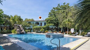 A swimming pool with a stone waterfall feature, surrounded by lush greenery and a building with a light blue facade.