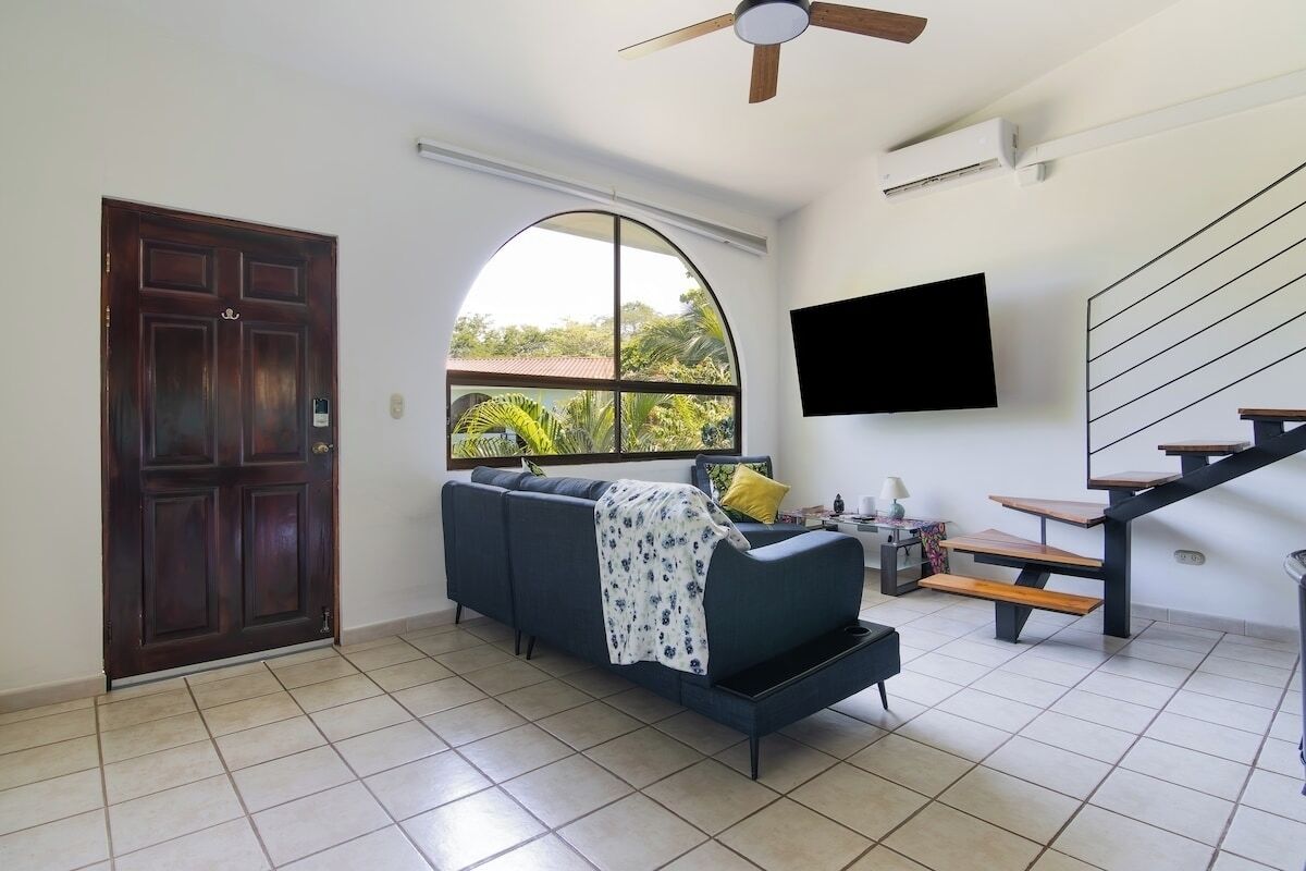 A living room with a dark-colored sofa, a TV mounted on the wall, a wooden staircase, and a large arched window.