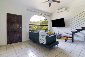 A living room with a dark-colored sofa, a TV mounted on the wall, a wooden staircase, and a large arched window.