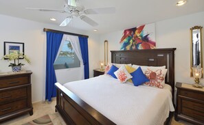Una camera da letto con un letto, una ventola a soffitto, un comodino con una lampada, una cassettiera, una finestra con tende e un quadro appeso al muro.