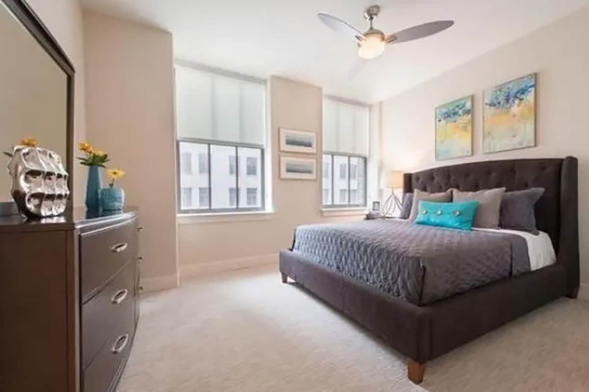 A bedroom with a bed, dresser, ceiling fan, and large windows.