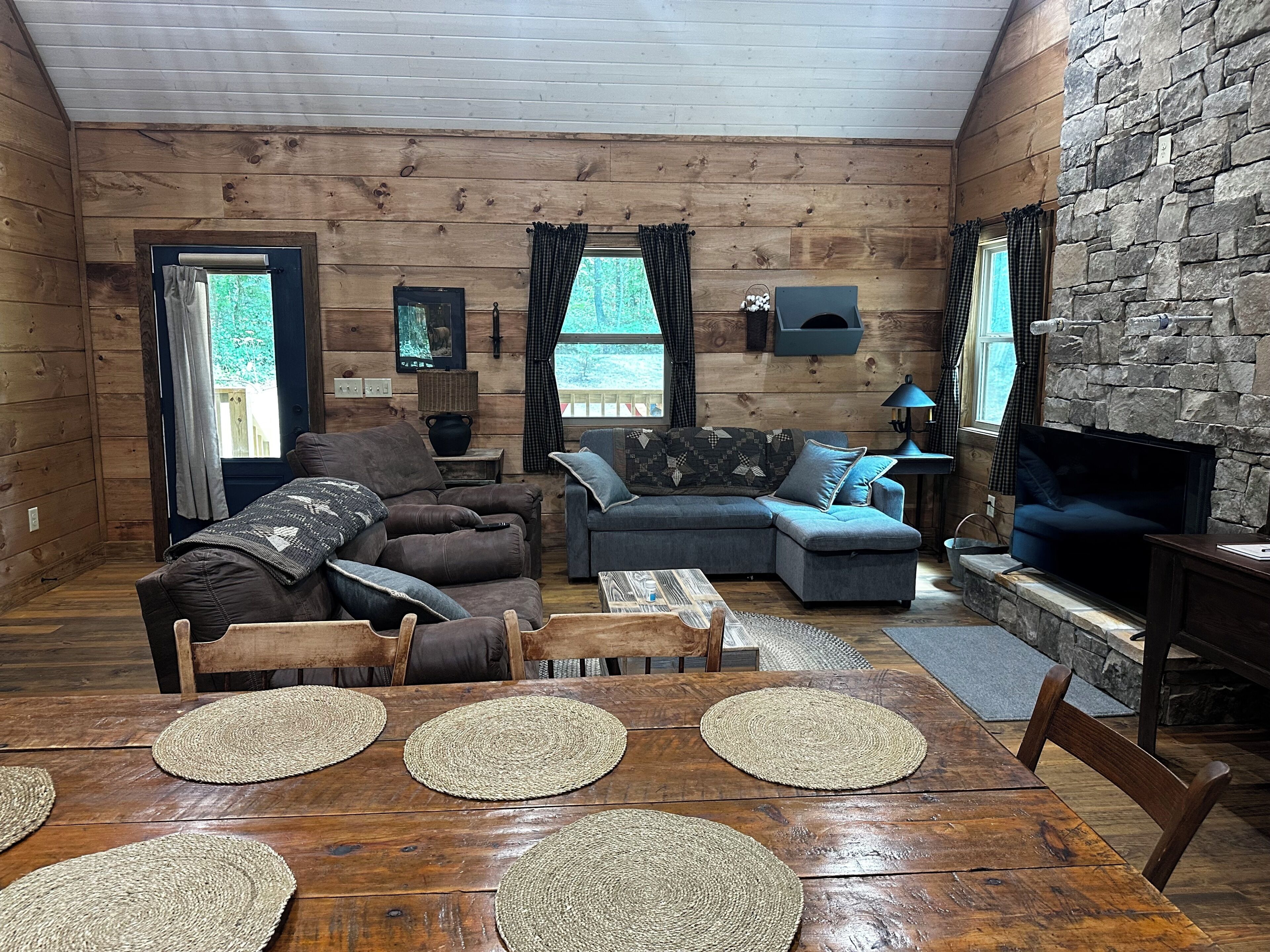 Interior acolhedor de uma cabana, com uma mesa de jantar de madeira, dois sofás, uma televisão e uma parede de pedra.