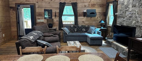 Interior acolhedor de uma cabana, com uma mesa de jantar de madeira, dois sofás, uma televisão e uma parede de pedra.