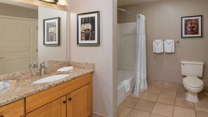 Un baño con encimera de granito, armarios de madera, un lavabo blanco, un inodoro, una ducha con cortina blanca y cuadros en la pared.