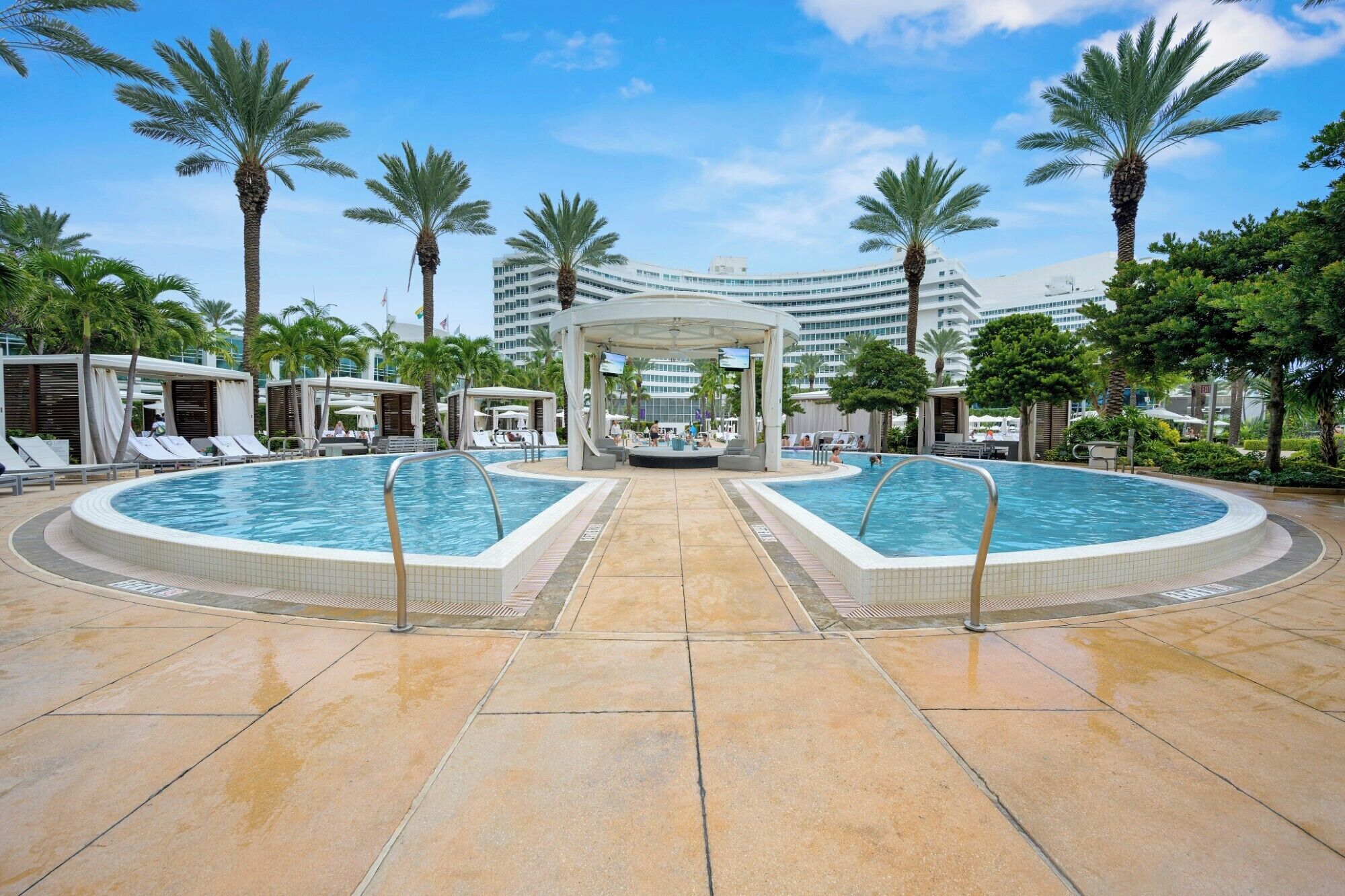 A resort pool area with two large pools, palm trees, and a covered seating area.