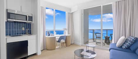 Una habitación de hotel moderna con cocina, zona de comedor, balcón con vistas al océano y un sofá azul.